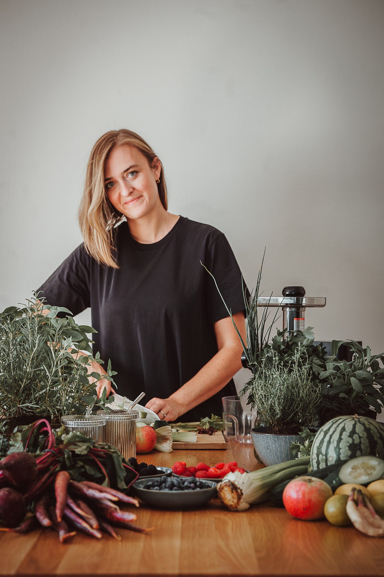 Stefanie Ensenberger steht lächelnd an einem Holztisch voller frischem Gemüse, Obst und Kräutern