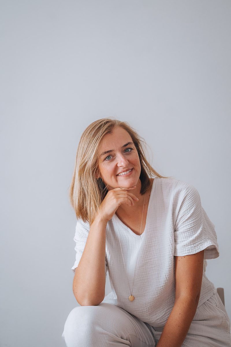 Stefanie Ensenberger sitzt in heller Kleidung entspannt vor einer hellen Wand und lächelt in die Kamera