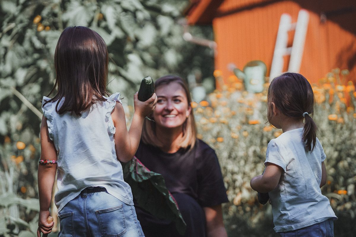 Stefanie Ensenberger mit ihren zwei Töchtern im Garten, eine Tochter reicht ihr eine Gurke
